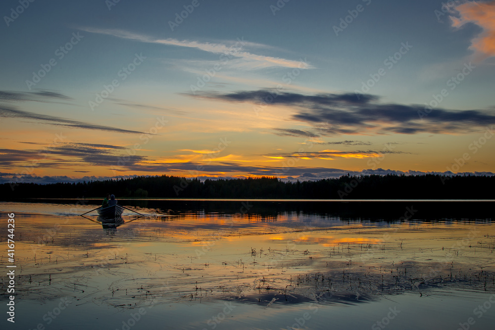 Two people are sailing in a boat. Beautiful sunset landscape over a Finnish lake. The space is flooded with gold.