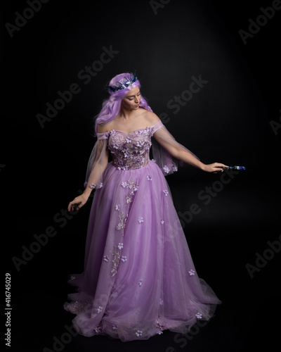 Full length portrait of girl wearing long purple fantasy ball gown with crown and pink hair, standing pose holding a wand against a studio background.