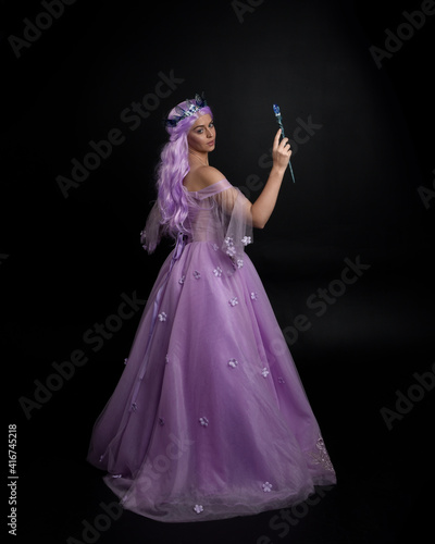 Full length portrait of girl wearing long purple fantasy ball gown with crown and pink hair  standing pose holding a wand  against a studio background.