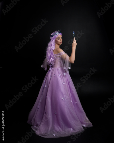 Full length portrait of girl wearing long purple fantasy ball gown with crown and pink hair, standing pose holding a wand against a studio background.