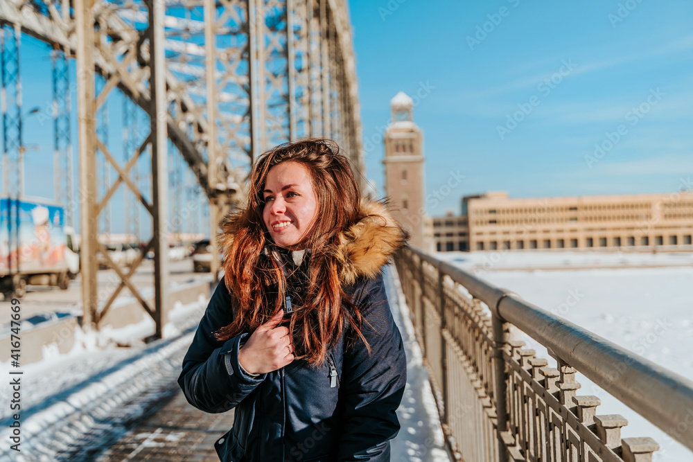 A young woman in a jacket walks on a snow bridge in St. Petersburg in winter in sunny weather