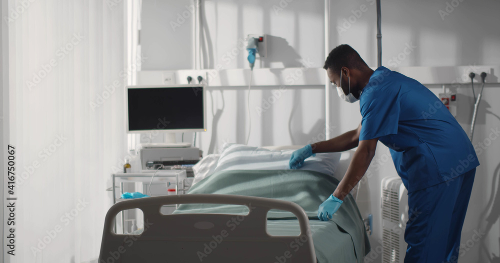 Fototapeta premium Expert young male nurse in mask working at hospital and making bed