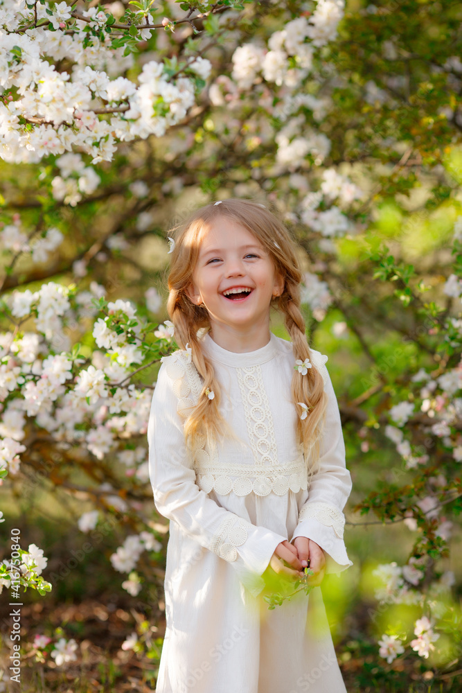 A sweet happy little girl in a white dress in spring near a flowering tree looks at the camera and laughs A sweet happy little girl in a white dress in spring near a flowering tree looks at the camera and laughs