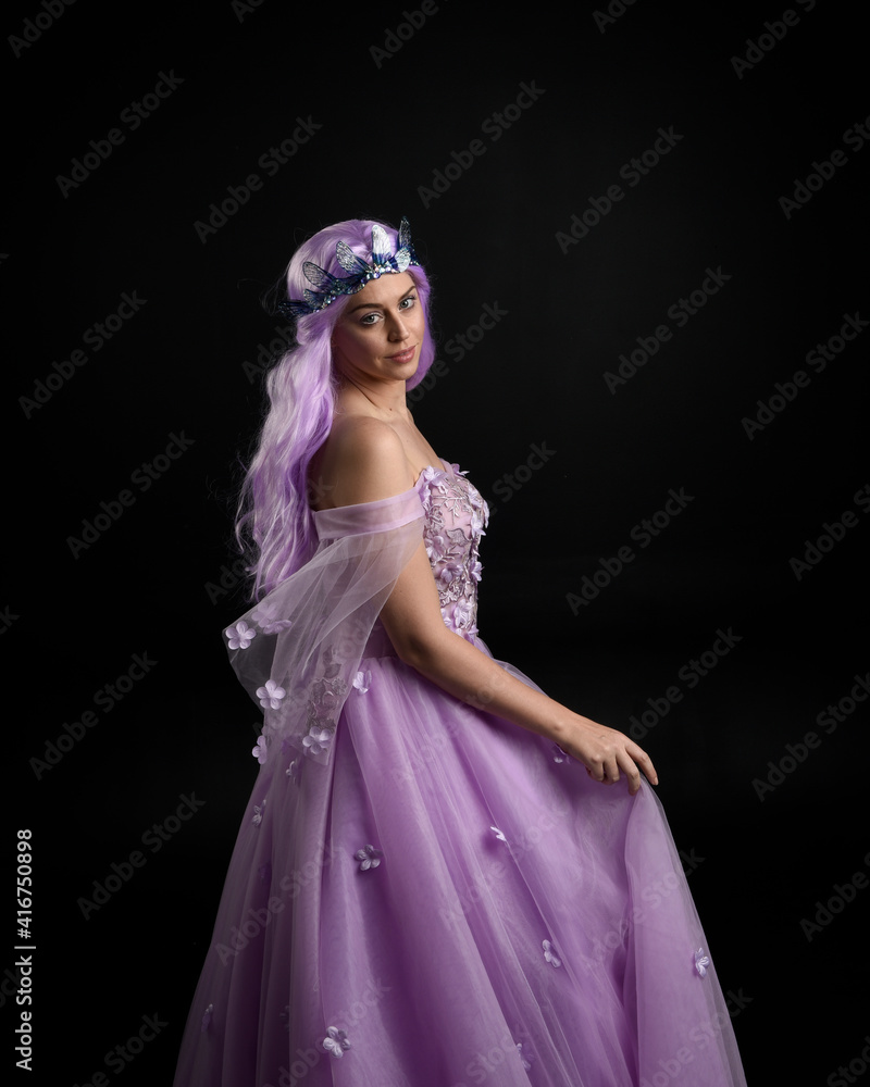 Close up portrait of girl wearing long purple fantasy ball gown with butterfly crown and pink hair, against a  dark studio background.
