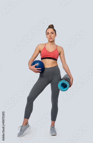 Fit young brunette girl exercise with blue medicine ball and gym mat on grey background