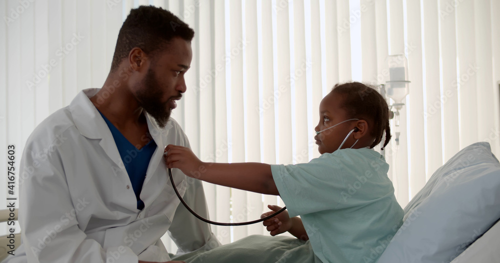 Obraz premium Cute african child playing doctor with man pediatrician on bed in hospital ward