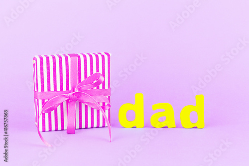 Happy Father's day. Gift box with a ribbon and a bow, the word "dad" from wooden letters on a light purple background. Copy space. Close-up.