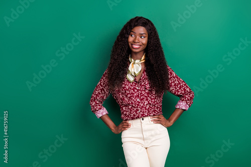 Photo of pretty cute curly dark skin woman dressed red shirt looking empty space arms waist isolated green color background © deagreez