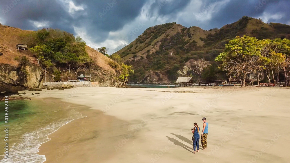 A couple walking on an idyllic Koka Beach. Hidden gem of Flores, Indonesia. Couple is enjoying their romantic escape. Waves gently washing the shore. There are hills in the back. Happiness and love A couple walking on an idyllic Koka Beach. Hidden gem of Flores, Indonesia. Couple is enjoying their romantic escape. Waves gently washing the shore. There are hills in the back. Happiness and love