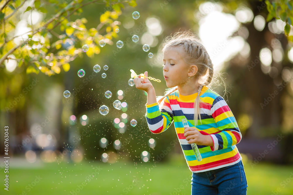 Cute little girl on a walk in a multi-colored knitted blouse in a striped summer blowing soap bubbles