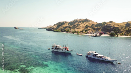 A drone shot of a paradise island with some boats anchored around in Komodo National Park, Flores, Indonesia. Brownish island turns into white sand beach and further into turquoise and navy sea.