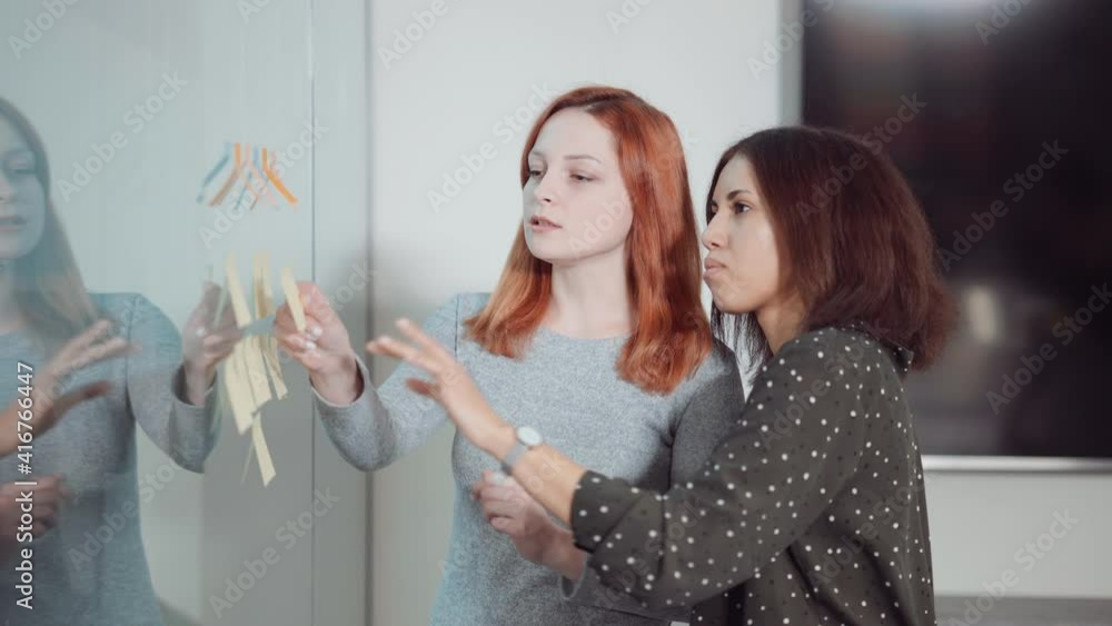 Multiethnic team of project managers sticking notes on Agile board arranged on glass wall in office. Business women checking tasks and controlling workflow. Concept of planning