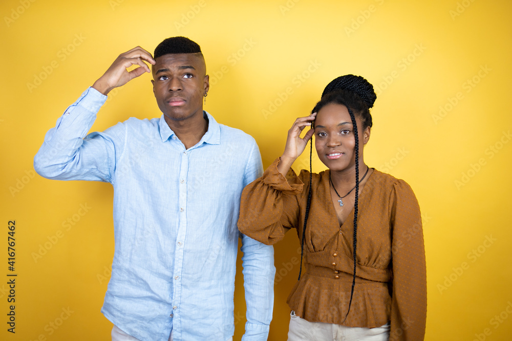 Young african american couple standing over yellow background onfuse and wonder about question. Uncertain with doubt, thinking with hand on head Young african american couple standing over yellow background onfuse and wonder about question. Uncertain with doubt, thinking with hand on head