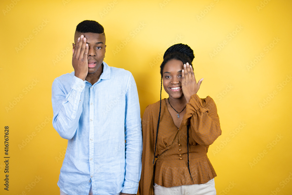 Young african american couple standing over yellow background covering one eye with hand, confident smile on face and surprise emotion. Young african american couple standing over yellow background covering one eye with hand, confident smile on face and surprise emotion.