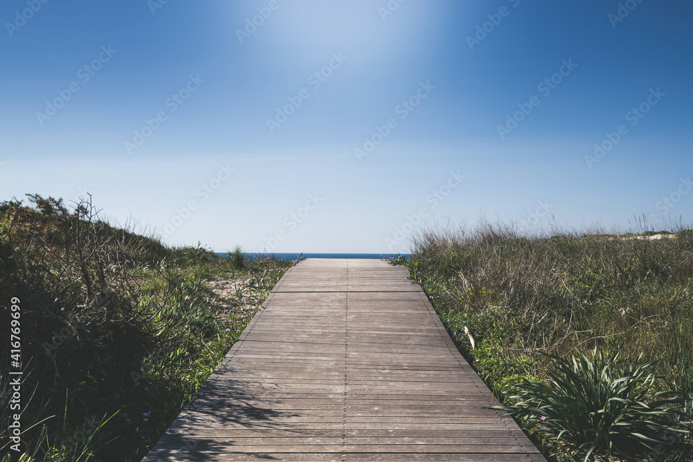 Close up view of wooden walkway leading to the sea through low Mediterranean vegetation. Path of wood that leads to infinity Blue sky in background. Positive concept of vacation freedom carefree peace