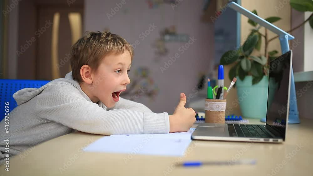 Excited little boy studying at home with laptop, showing thumb up to the laptop screen. Training books and notebook on the table. Distance learning online education Excited little boy studying at home with laptop, showing thumb up to the laptop screen. Training books and notebook on the table. Distance learning online education
