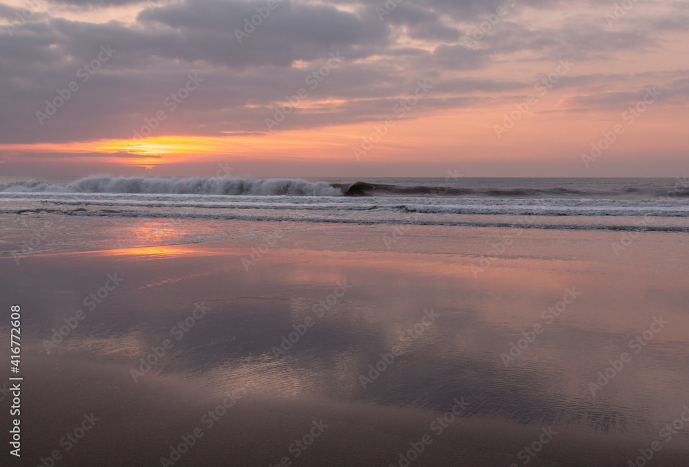 Waves crash against Welcombe Mouth beach in north Devon during sunset. The sunset is reflected in the sand.