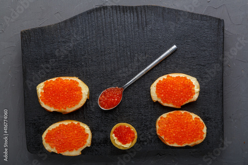Canapes with butter and red caviar and a tartlet and a spoon with caviar on a black board on a concrete background.