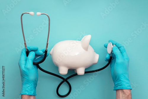 Health care cost. Doctor in surgical gloves holding a white piggy bank and stethoscope photo