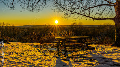 Granby Connecticut.  A picnic table on a mountain top rests in snow and mud, while illuminated by a sunset view of another mountain scene.   Beautiful sunset winter scene.