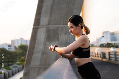 Selective focus at face of young beautiful Asian women using smart watch to track activity and listen music while warm up before exercise or running at the urban city view. Fit and healthy lifestyle.