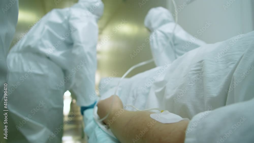 Close up shot of hand of emergency patient with IV drip being transported on gurney by paramedics in protective suits through hospital hallway Close up shot of hand of emergency patient with IV drip being transported on gurney by paramedics in protective suits through hospital hallway