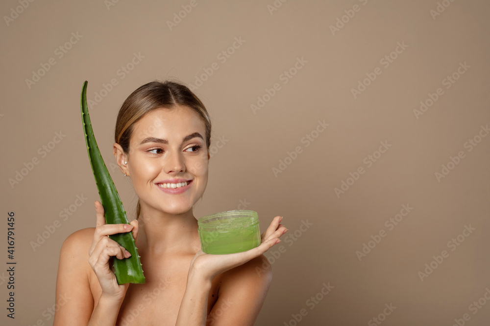 Smiling happy young female model holding aloe leaf and jar of aloe gel on beige background. The concept of skin care, moisturizing with natural cosmetics Smiling happy young female model holding aloe leaf and jar of aloe gel on beige background. The concept of skin care, moisturizing with natural cosmetics