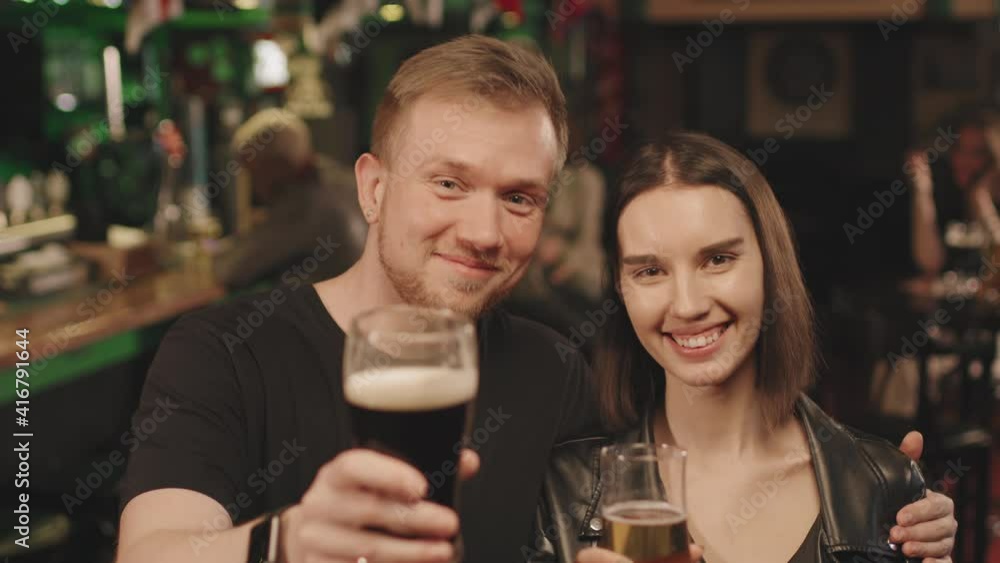 Medium close-up portrait with slowmo of smiling young couple looking at camera holding glasses of beer having fun together in local bar Medium close-up portrait with slowmo of smiling young couple looking at camera holding glasses of beer having fun together in local bar