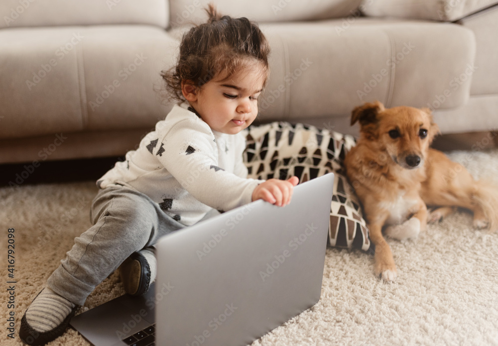 Adorable toddler sitting on the floor touching laptop and near a dog during the covid-19 pandemic lock down