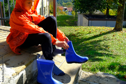 Boy in orange raincoat putting on blue rubby boots sitting on stairs in backyard. Clothing for rainy weather	
