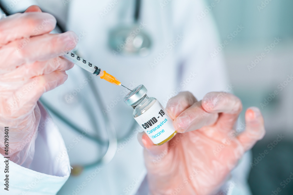 Doctor or nurse in uniform and gloves wearing face mask protective in lab holding medicine vial vaccine bottle with COVID-19 Coronovirus vaccine label Doctor or nurse in uniform and gloves wearing face mask protective in lab holding medicine vial vaccine bottle with COVID-19 Coronovirus vaccine label