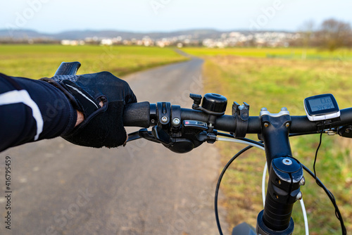 A bicycle handlebar seen from the first person perspective and with a mans hand on the handle. Visible bike computer and bell.