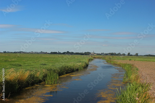 Wide view over Dutch meadows and a small river with cows grazing in the distance and a village with a church. Photo was taken on a sunny day with a blue sky.