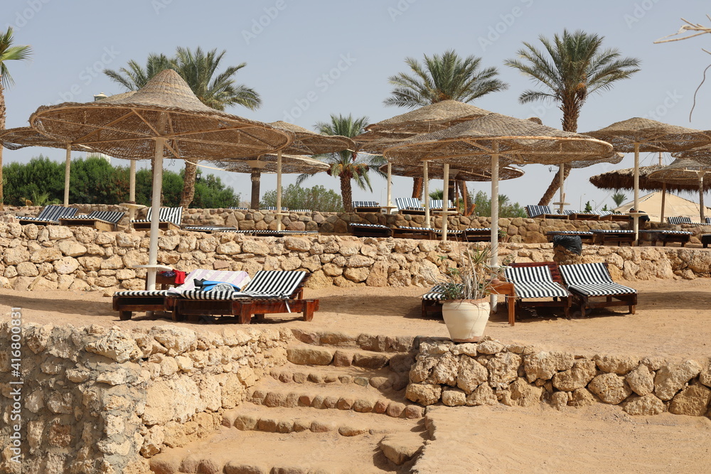 Naklejka premium Scenic sunny landscape. Luxury beach with deck chair in the evening time. Beautiful view of Red Sea coast. Comfortable beach with lounge chairs under natural wooden umbrella