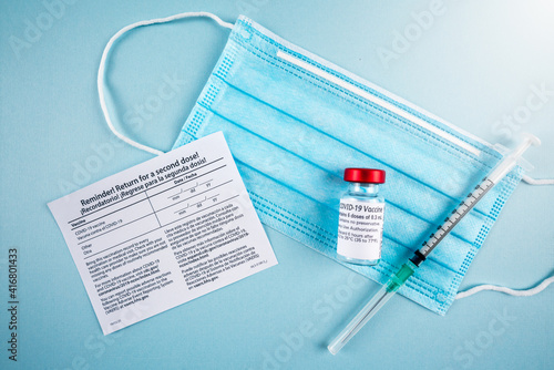 return for a second dose card with facial mask, covid-19 vaccine vial and syringe on blue background.