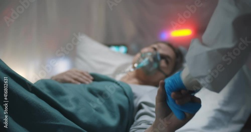 Doctor in gloves and ppe suit holding hands of sick covid-19 aged lady lying in bed
