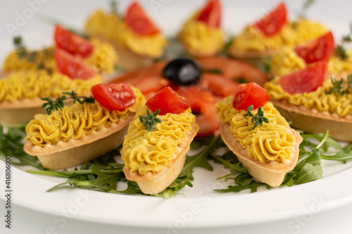 Blurred image of pate tartlets decorated with tomato slices on a white plate with herbs.