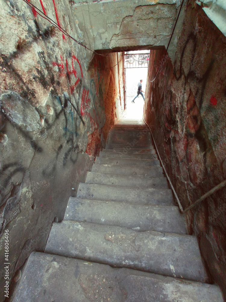 Going down some old stairs in an abandoned building with graffiti painted walls, in the background a man is walking along the sidewalk.