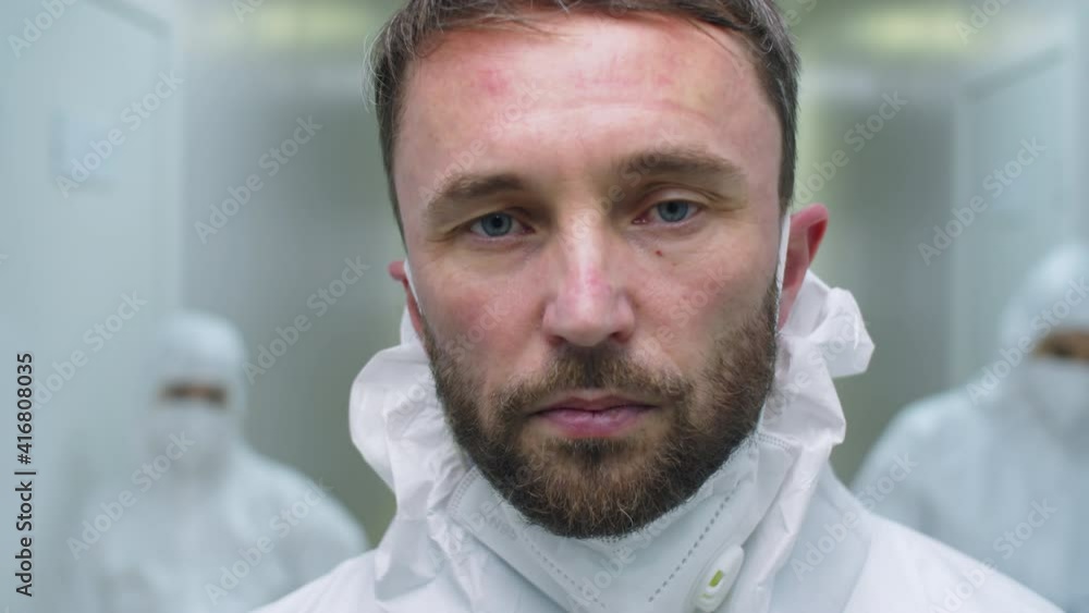 Portrait of tired male doctor in protective uniform and mask under chin standing in hospital and looking at camera while working in time of covid-19 pandemic Portrait of tired male doctor in protective uniform and mask under chin standing in hospital and looking at camera while working in time of covid-19 pandemic