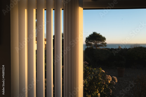 Half open vertical blinds with a golden glow as the sun sets.There is a tree and a view of the sea through the window.