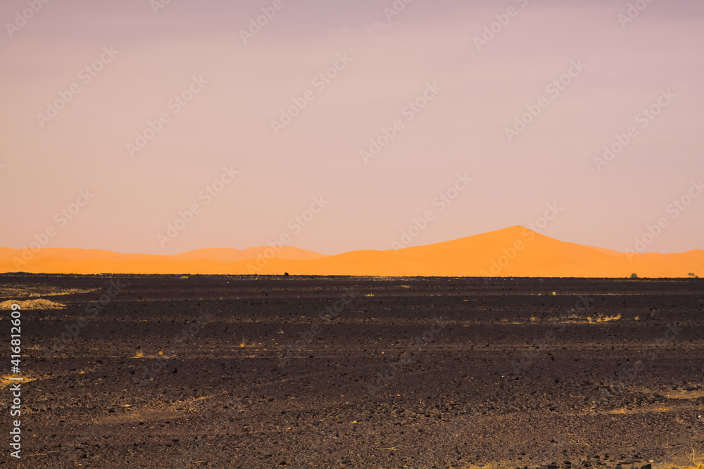 View over endless black flat waste scorched earth on golden yellow sand dunes with blurred gloomy sky in the dawn of morning, Erg Chebbi, Morocco View over endless black flat waste scorched earth on golden yellow sand dunes with blurred gloomy sky in the dawn of morning, Erg Chebbi, Morocco