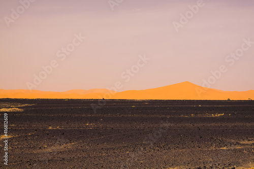 View over endless black flat waste scorched earth on golden yellow sand dunes with blurred gloomy sky in the dawn of morning, Erg Chebbi, Morocco