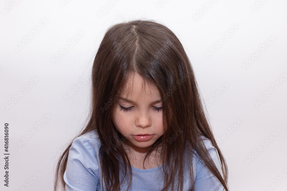Beautiful little cute girl with long brown even hair looks sadly down on a white background in a studio Beautiful little cute girl with long brown even hair looks sadly down on a white background in a studio