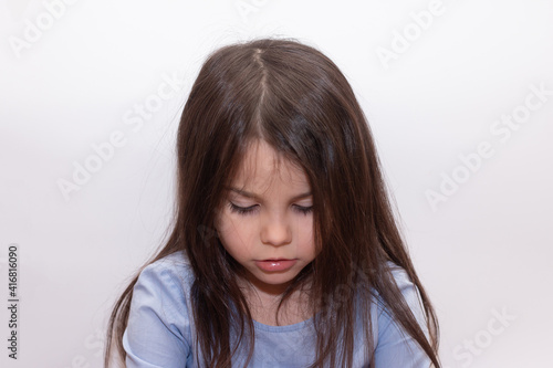 Beautiful little cute girl with long brown even hair looks sadly down on a white background in a studio