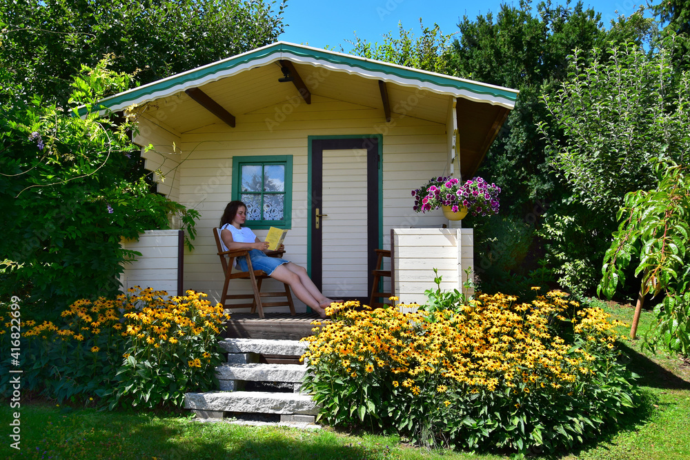 A teenage girl sitting in a chair on the porch of a garden shed, reading a book.