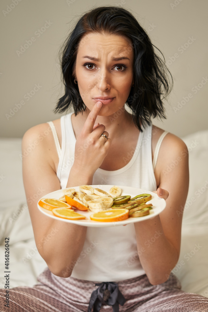 a young woman with shoulder-length dark hair in light-colored pajamas looks at a plate of sliced fruits and frowns, pressing her finger to her lips. a young woman with shoulder-length dark hair in light-colored pajamas looks at a plate of sliced fruits and frowns, pressing her finger to her lips.