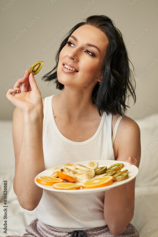 a beautiful brunette girl in pajamas holds a plate with sliced fruits in her hands and a slice of kiwi in her other hand and looks dreamily to the side and smiles a beautiful brunette girl in pajamas holds a plate with sliced fruits in her hands and a slice of kiwi in her other hand and looks dreamily to the side and smiles