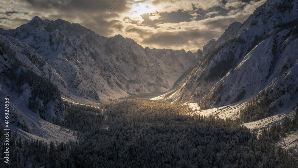 Aerial flight over the mountain valley in winter covered in snow. Cinematic drone view with sunlight breaking through clouds.
