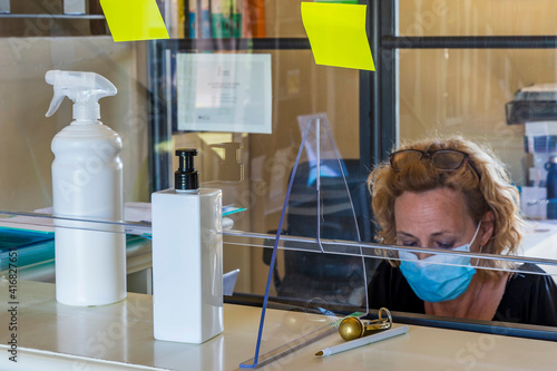 A woman with a protective mask works in an office at the reception of a hotel separated by a sheet of plexiglass photo