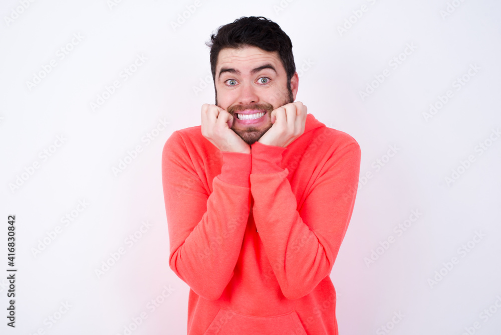 Dreamy young Caucasian bearded man wearing pink hoodie against white background keeps hands pressed together under chin, looks with happy expression, has toothy smile. Dreamy young Caucasian bearded man wearing pink hoodie against white background keeps hands pressed together under chin, looks with happy expression, has toothy smile.